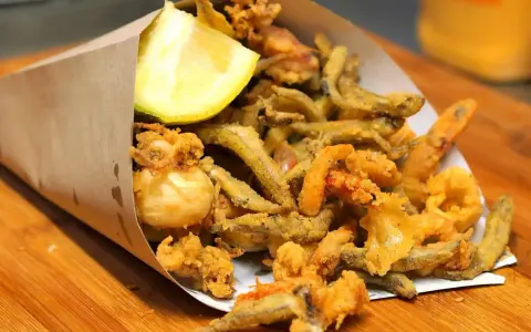 Un cono di carta marrone traboccante di una deliziosa frittura di pesce croccante, con calamari, gamberetti e pesciolini, guarnito con uno spicchio di limone.