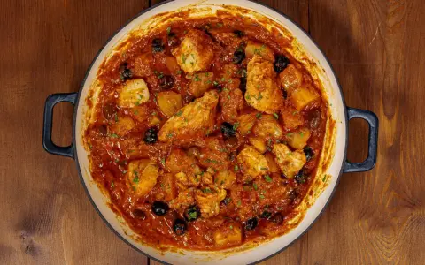 Un saporito baccalà in umido con patate e olive nere, guarnito con prezzemolo fresco, in una casseruola su un tavolo di legno.