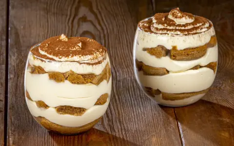 Due bicchieri di delizioso tiramisù senza uova, con strati di crema e savoiardi, spolverati di cacao.
