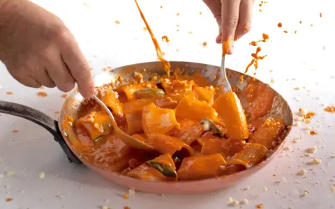 Mani che mescolano paccheri alla Vittorio in padella, con salsa arancione e schizzi di sugo.