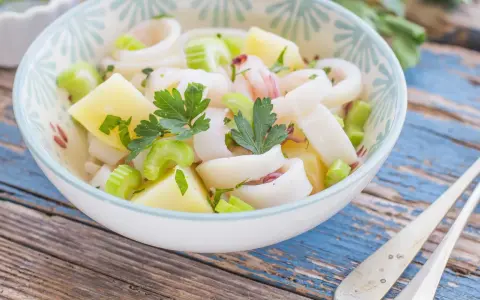Insalata di calamari fresca con patate, sedano e prezzemolo, servita in una ciotola decorata su un tavolo di legno blu.