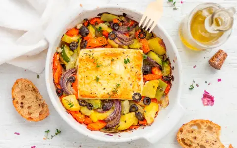 Feta al forno con verdure miste, olive nere e erbe aromatiche, accompagnata da pane rustico e olio d'oliva.
