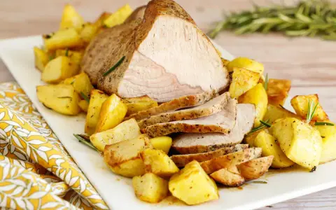 Un succulento arrosto di vitello affettato, servito su un piatto bianco con patate arrosto dorate e rametti di rosmarino.
