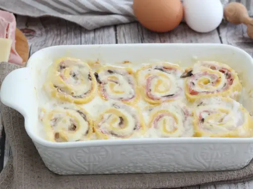 Rotolini di pasta con prosciutto e formaggio, cotti in una cremosa besciamella, sembrano piccoli nidi di rondine pronti per essere gustati.