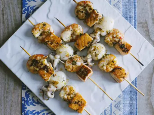 Spiedini di pesce misti, con gamberi, calamari e salmone, conditi con erbe aromatiche e serviti su un piatto bianco.