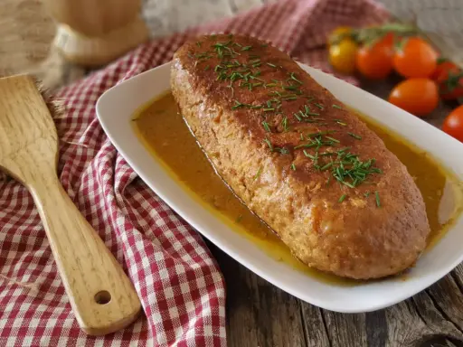 Polpettone in padella dorato e succulento, guarnito con erba cipollina, servito su un piatto bianco con salsa. Accanto, una spatola in legno e pomodorini.