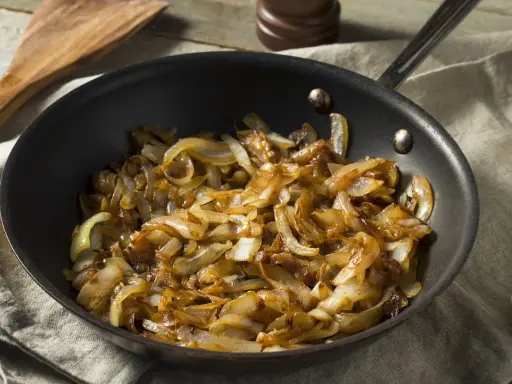 Cipolle caramellate in padella, pronte per insaporire un piatto. Un cucchiaio di legno attende accanto.