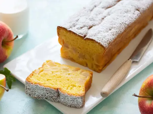 Un delizioso plumcake alle mele, cosparso di zucchero a velo, con una fetta tagliata e mele rosse fresche accanto.