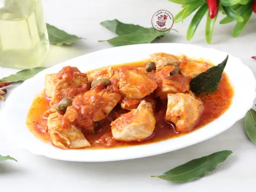 Succulento pollo alla pizzaiola in un piatto bianco, con capperi, pomodoro e foglie di alloro. Accanto, peperoncini secchi e una bottiglia d'olio.