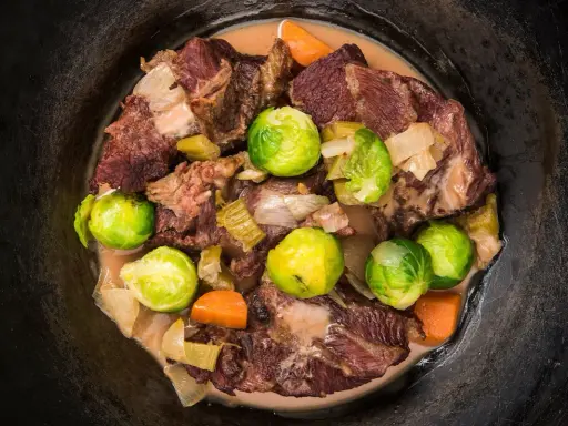 Guancia di vitello stufata con cavolini di Bruxelles, carote e cipolle in un pentolone scuro.