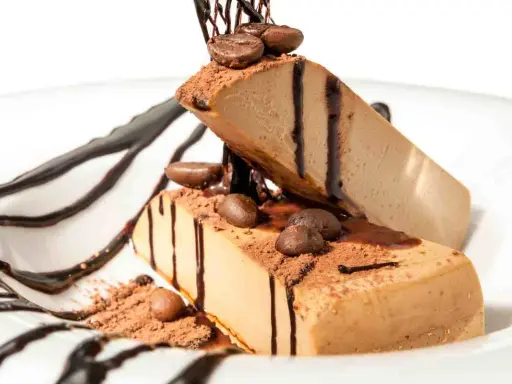 Due fette di semifreddo al caffè, decorate con cacao, chicchi di caffè e salsa al cioccolato, su un piatto bianco.