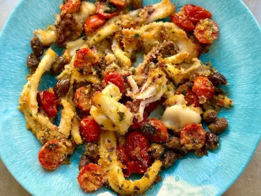 Calamari gratinati al forno su piatto blu, con pomodorini e olive. Croccanti e saporiti, un piatto che profuma di mare.