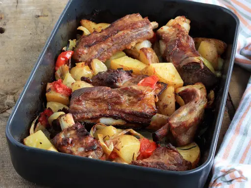 Succulente costine al forno con patate, peperoni e cipolle in una teglia scura.