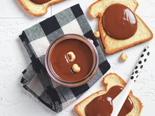 Un vasetto di crema di nocciole fatta in casa, con nocciole intere, su pane tostato e tovagliolo a scacchi.