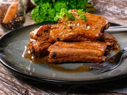 Succulente costine di maiale glassate, guarnite con prezzemolo fresco, servite su un piatto scuro con una forchetta.