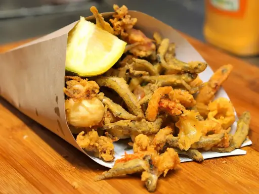 Un cono di carta marrone traboccante di una deliziosa frittura di pesce croccante, con calamari, gamberetti e pesciolini, guarnito con uno spicchio di limone.