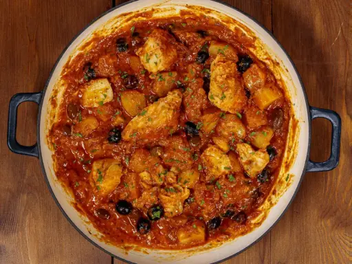 Un saporito baccalà in umido con patate e olive nere, guarnito con prezzemolo fresco, in una casseruola su un tavolo di legno.