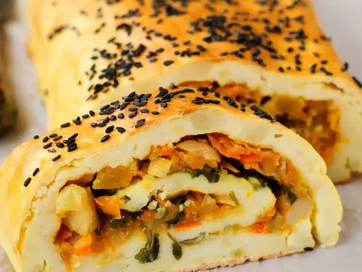 Rotolo di patate dorato, cosparso di semi neri, con un ripieno colorato di verdure.