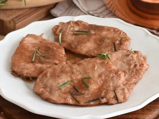 Quattro scaloppine di maiale succulente, guarnite con rosmarino fresco, servite su un piatto bianco.