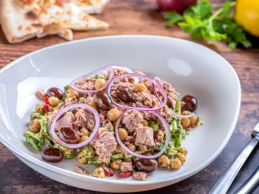 Insalata di ceci e tonno con olive nere e cipolla rossa, servita in un piatto bianco su un tavolo di legno.