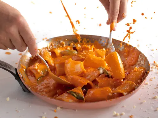 Mani che mescolano paccheri alla Vittorio in padella, con salsa arancione e schizzi di sugo.