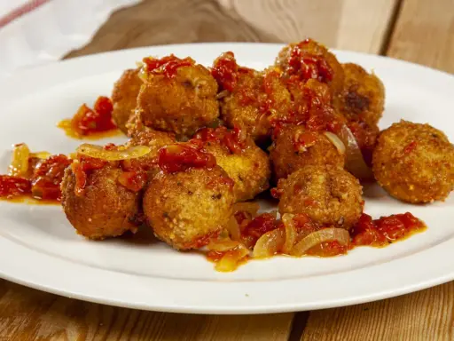 Polpette di pesce dorate e saporite, condite con un sugo rosso di pomodoro e cipolla, servite su un piatto bianco.