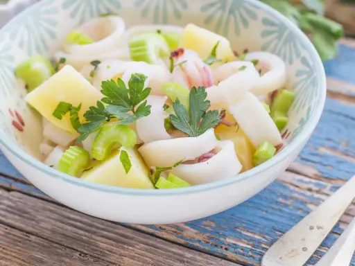 Insalata di calamari fresca con patate, sedano e prezzemolo, servita in una ciotola decorata su un tavolo di legno blu.