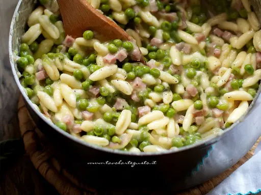 Un primo piatto invitante: pasta e piselli cremosa con cubetti di prosciutto cotto, mescolata con un cucchiaio di legno.