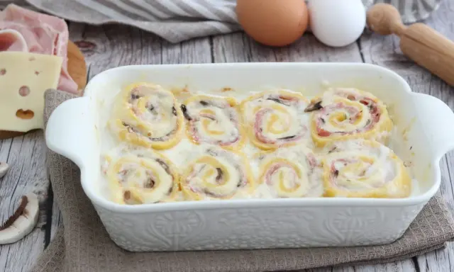 Rotolini di pasta con prosciutto e formaggio, cotti in una cremosa besciamella, sembrano piccoli nidi di rondine pronti per essere gustati.