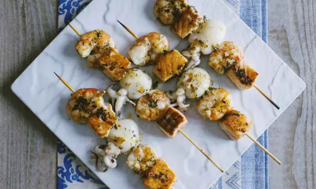 Spiedini di pesce misti, con gamberi, calamari e salmone, conditi con erbe aromatiche e serviti su un piatto bianco.