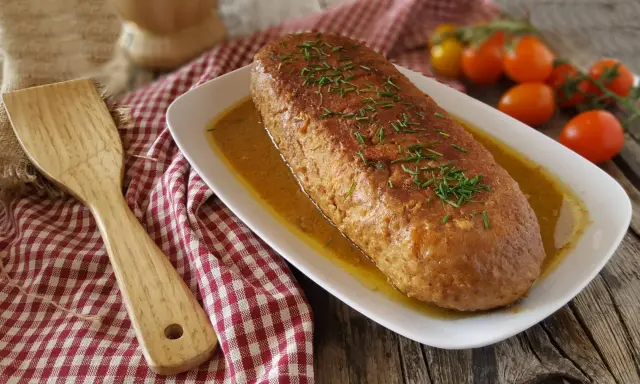 Polpettone in padella dorato e succulento, guarnito con erba cipollina, servito su un piatto bianco con salsa. Accanto, una spatola in legno e pomodorini.