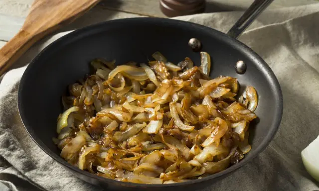 Cipolle caramellate in padella, pronte per insaporire un piatto. Un cucchiaio di legno attende accanto.