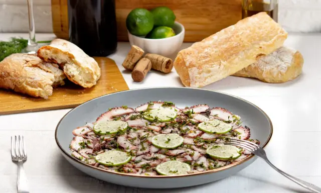 Delizioso carpaccio di polpo, guarnito con fette di lime e erbe aromatiche, pronto per essere gustato con pane fresco e vino.