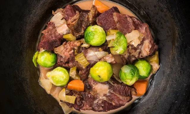 Guancia di vitello stufata con cavolini di Bruxelles, carote e cipolle in un pentolone scuro.