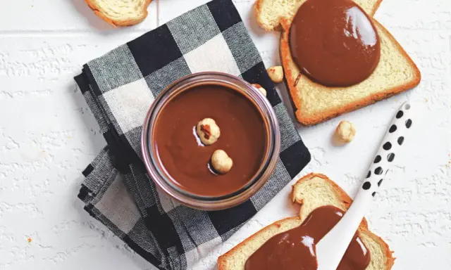 Un vasetto di crema di nocciole fatta in casa, con nocciole intere, su pane tostato e tovagliolo a scacchi.