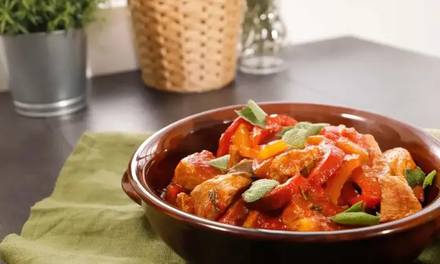 Un saporito spezzatino di vitello con peperoni rossi e gialli, guarnito con foglie di salvia fresca, servito in una ciotola di terracotta.
