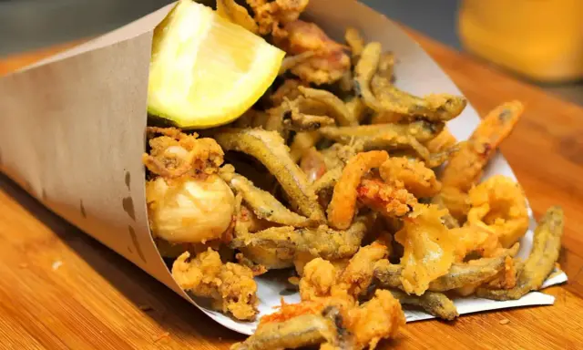 Un cono di carta marrone traboccante di una deliziosa frittura di pesce croccante, con calamari, gamberetti e pesciolini, guarnito con uno spicchio di limone.