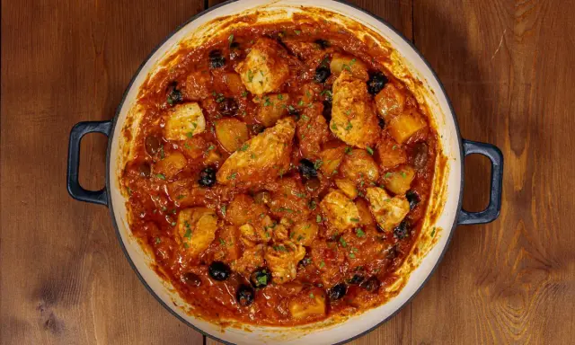 Un saporito baccalà in umido con patate e olive nere, guarnito con prezzemolo fresco, in una casseruola su un tavolo di legno.