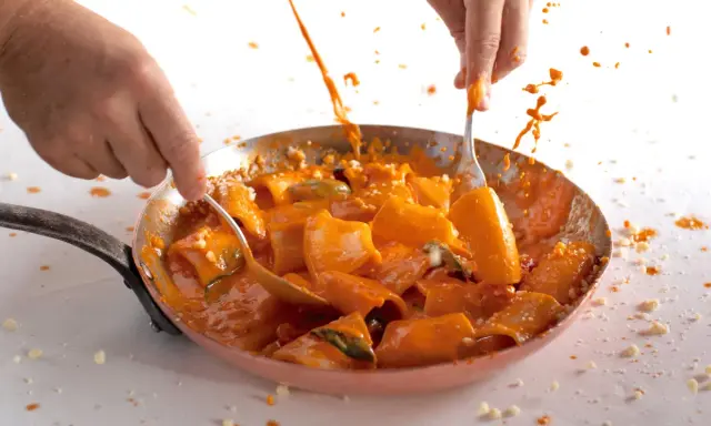 Mani che mescolano paccheri alla Vittorio in padella, con salsa arancione e schizzi di sugo.