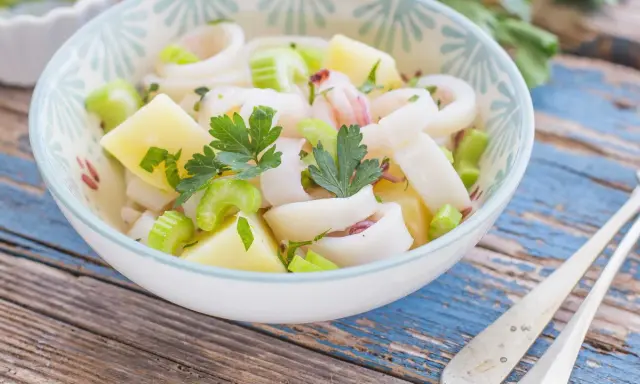 Insalata di calamari fresca con patate, sedano e prezzemolo, servita in una ciotola decorata su un tavolo di legno blu.