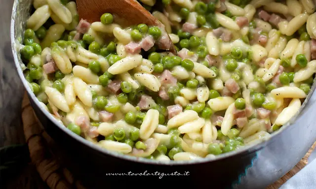 Un primo piatto invitante: pasta e piselli cremosa con cubetti di prosciutto cotto, mescolata con un cucchiaio di legno.