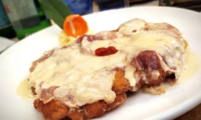 Cotoletta alla bolognese dorata e filante, con prosciutto e formaggio fuso, servita su un piatto bianco con contorno di purè e carota.