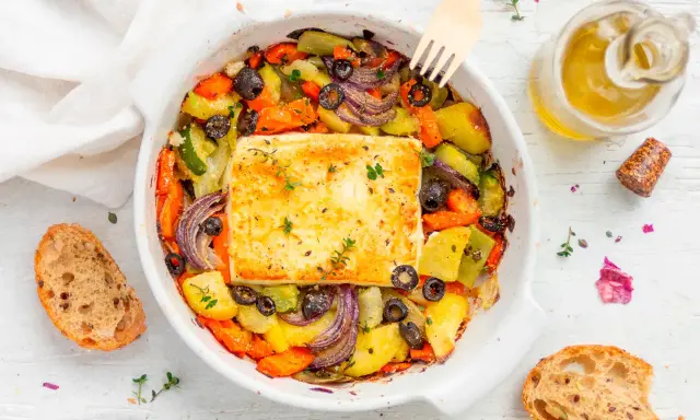 Feta al forno con verdure miste, olive nere e erbe aromatiche, accompagnata da pane rustico e olio d'oliva.