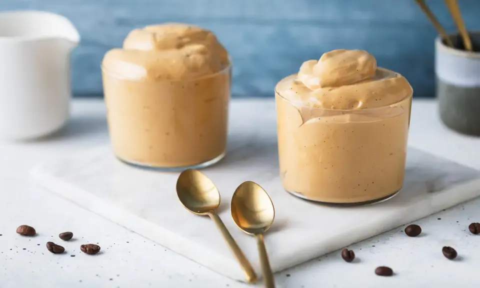 Due bicchieri di cremosa schiuma di caffè, perfetta per una ricetta crema di caffè senza panna. Due cucchiaini dorati e chicchi di caffè completano la scena.