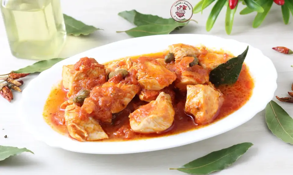 Succulento pollo alla pizzaiola in un piatto bianco, con capperi, pomodoro e foglie di alloro. Accanto, peperoncini secchi e una bottiglia d'olio.