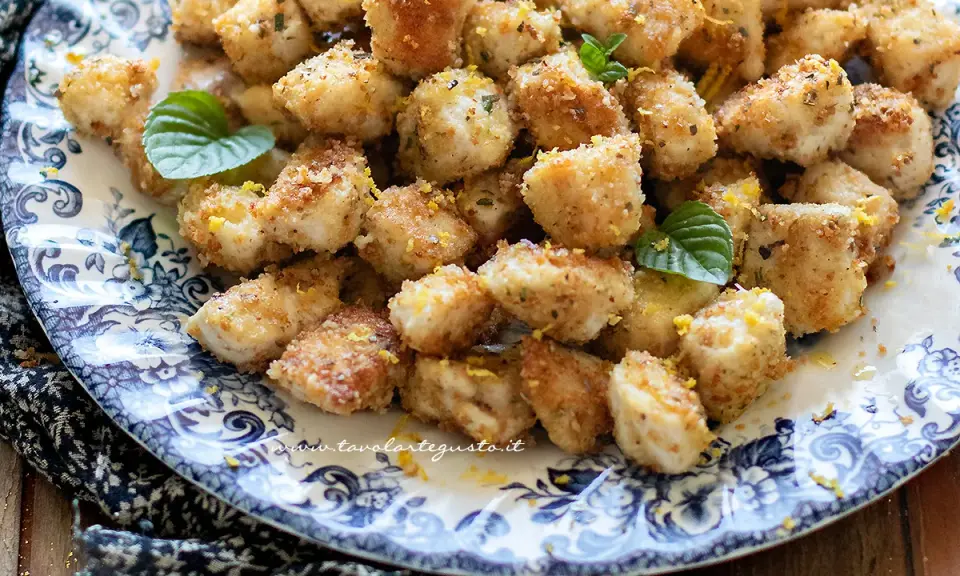 Bocconcini di pollo dorati e croccanti, guarniti con scorza di limone e foglioline di menta, serviti su un piatto decorato.