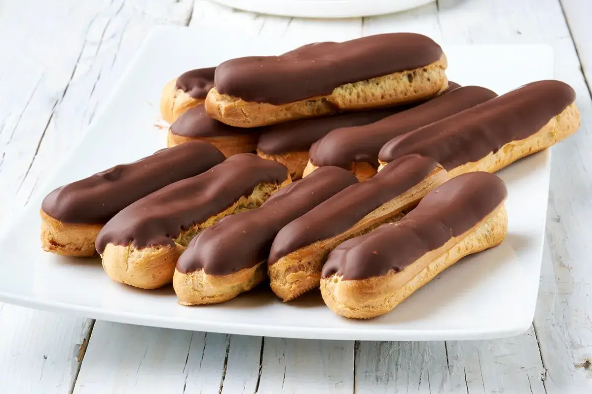 Un piatto bianco pieno di deliziosi dolci al cioccolato, eclair ricoperti di glassa fondente su una superficie di legno chiaro.