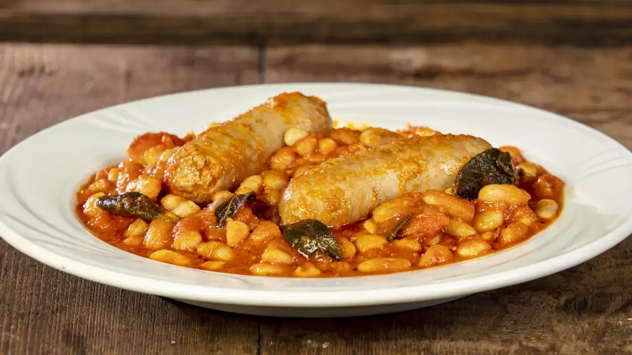 Piatto di salsiccia e fagioli in umido, con pomodoro e erbe aromatiche, servito su un piatto bianco.