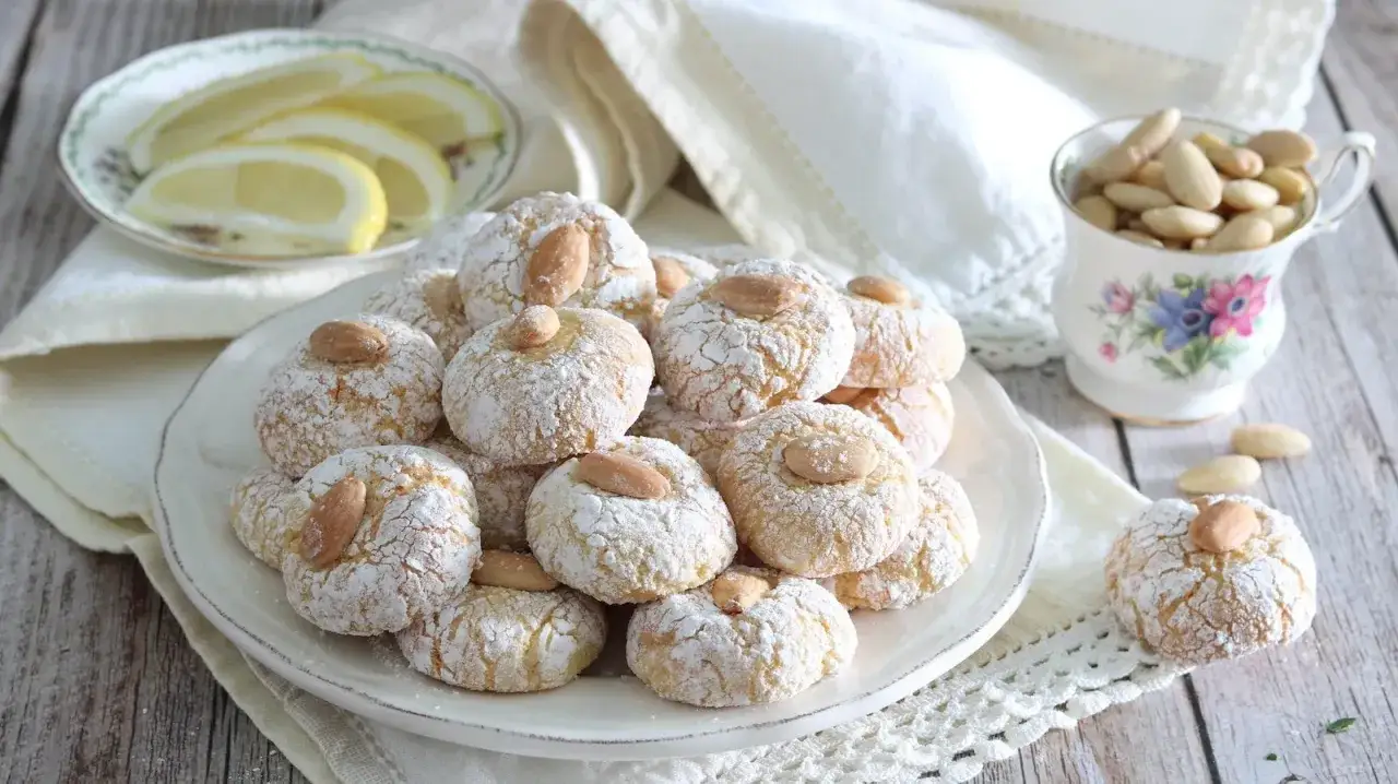 Biscotti con farina di mandorle, spolverati di zucchero a velo e guarniti con una mandorla intera. Accanto, spicchi di limone e una tazza piena di mandorle.