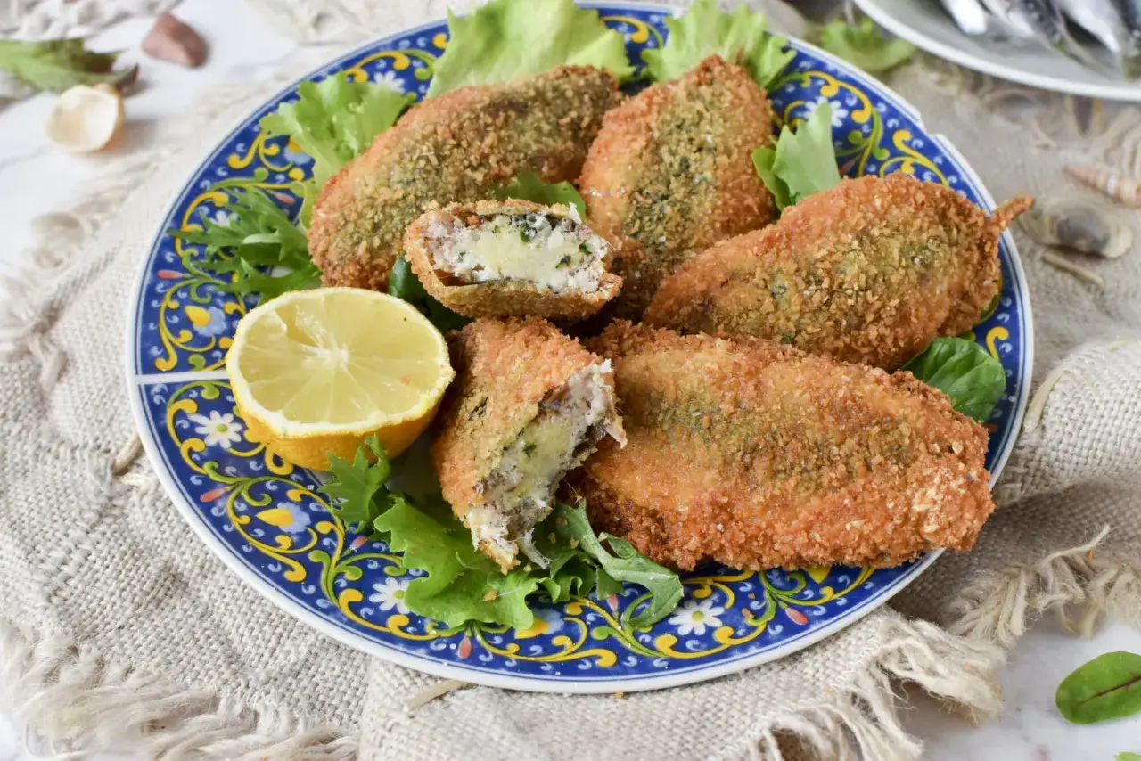Sarde a beccafico dorate e croccanti, ripiene e servite su un piatto blu decorato con foglie verdi e spicchio di limone.